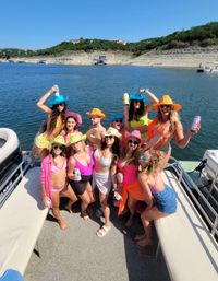Group of friends on a pontoon boat lake party in colorful swimsuits and neon cowboy hats, holding drinks with rocky shoreline and lakeside homes in the background