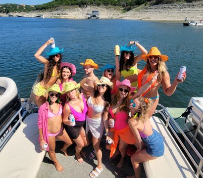 Group of friends on a pontoon boat lake party in colorful swimsuits and neon cowboy hats, holding drinks with rocky shoreline and lakeside homes in the background