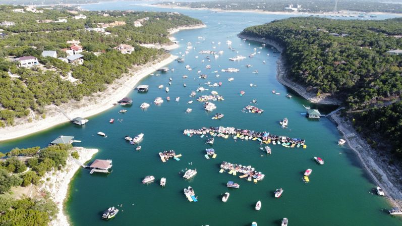 Aerial view of a busy lake cove with dozens of boats and colorful floating platforms clustered near tree-lined limestone shores on a sunny day
