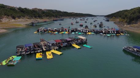 Aerial drone view of a summer lake raft-up with dozens of pontoon boats linked together, colorful floating mats and sunbathers near a tree-lined shoreline