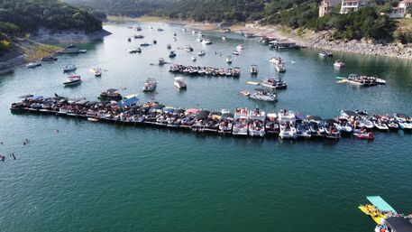 Aerial view of a crowded lake party with dozens of pontoon and motorboats rafted together, swimmers in the water, and wooded shoreline on a sunny summer day