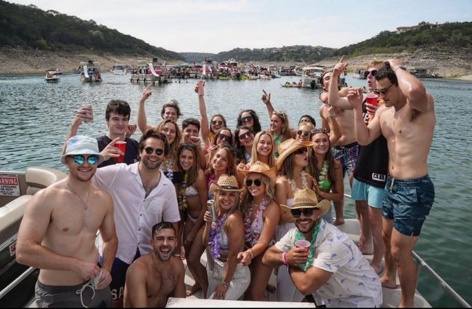 Energetic summer pontoon boat party on a crowded lake with a large group of friends in swimsuits, straw hats and red cups, surrounded by other boats and rocky shoreline