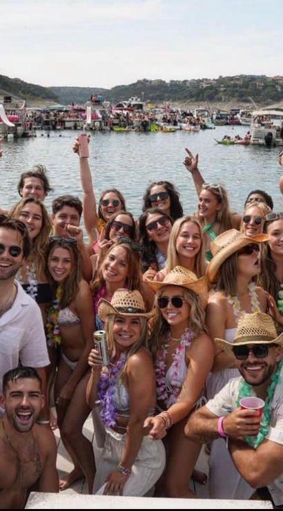 Energetic summer pontoon boat party on a crowded lake with a large group of friends in swimsuits, straw hats and red cups, surrounded by other boats and rocky shoreline
