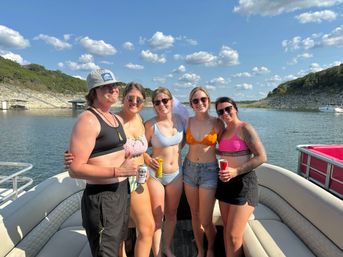 Five friends in swimsuits smiling on a pontoon boat at a sunny lake with rocky shorelines and puffy clouds, holding canned drinks on a summer boating day.