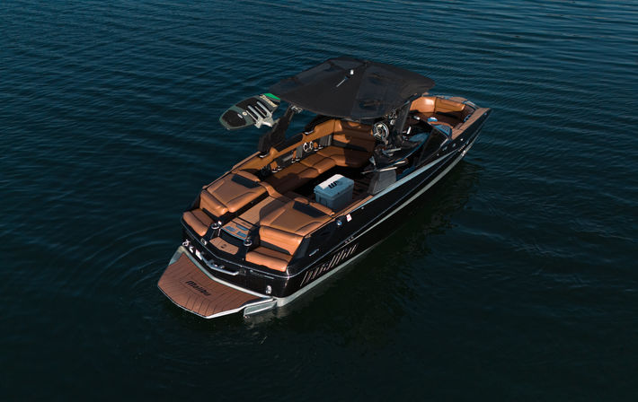Aerial view of a luxury black wakeboat with tan leather seating, wakeboard tower and swim platform floating on calm deep-blue lake water.