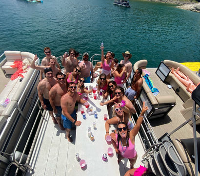 Aerial view of a large group celebrating on a pontoon boat at a sunny lake, friends in swimsuits holding drinks and cheering