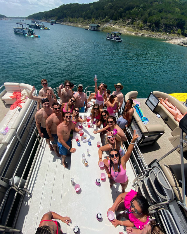 Aerial view of a large group celebrating on a pontoon boat at a sunny lake, friends in swimsuits holding drinks and cheering