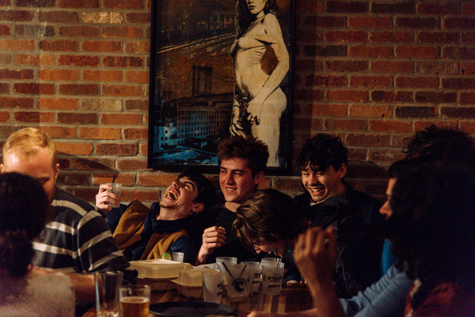 Group of young people laughing together at a crowded table in a cozy brick-walled bar, drinks and takeout containers on the table and a framed vintage-style poster on the wall — lively night out.