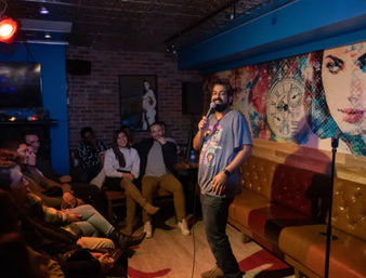 Comedian performing stand-up in a cozy comedy club, holding a microphone as a seated audience laughs by a brick wall and colorful mural.