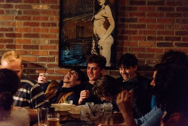 Group of friends laughing around a cluttered table with drinks and takeout in a cozy brick‑walled bar beneath framed artwork of a woman.