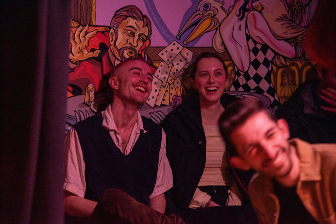 Three people laughing in a dimly lit live-show venue, seated in front of a colorful mural of playing cards and a stylized bird — lively nightlife atmosphere.