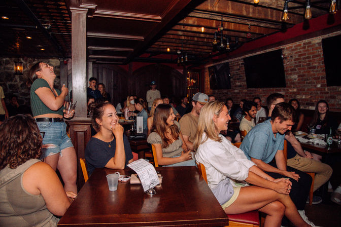 Lively crowd of young adults laughing during a comedy night at a cozy brick‑walled pub with wooden beams, Edison bulbs, drinks and menus on tables.