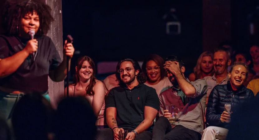 Comedian onstage with a microphone as a smiling, drink-holding audience laughs at an intimate live comedy club show