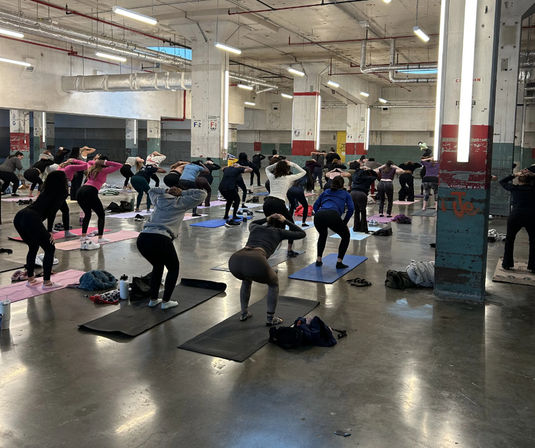 Group fitness class in a converted industrial warehouse — dozens doing squats on yoga mats amid concrete floors, exposed pipes and pillars, with workout bags and water bottles scattered around.