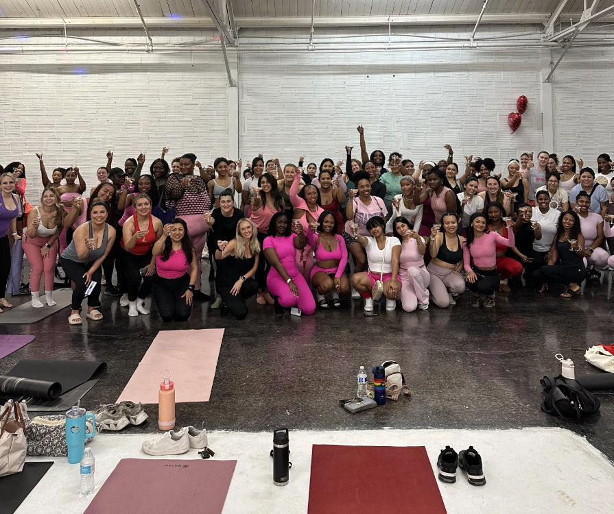 Large group of women in pink activewear posing and smiling in a bright indoor fitness studio for a group-fitness celebration, with yoga mats, water bottles and workout gear in the foreground