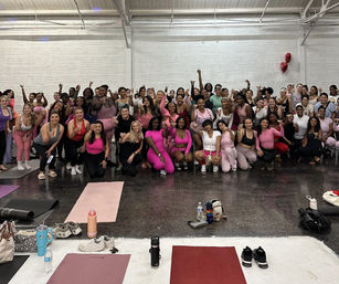 Large group of women in pink activewear posing and smiling in a bright indoor fitness studio for a group-fitness celebration, with yoga mats, water bottles and workout gear in the foreground
