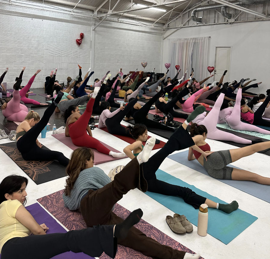 Large group fitness class in a bright industrial studio, participants on mats doing side leg‑lift Pilates/yoga stretches in colorful leggings with heart-shaped balloons overhead