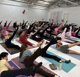 Large group fitness class in a bright industrial studio, participants on mats doing side leg‑lift Pilates/yoga stretches in colorful leggings with heart-shaped balloons overhead