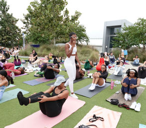 Outdoor group yoga/fitness class on a grassy lawn by the waterfront, instructor in white guides participants on mats through seated core poses.
