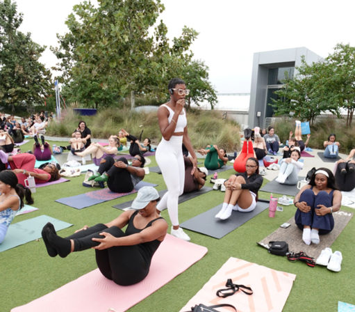 Outdoor group yoga/fitness class on a grassy lawn by the waterfront, instructor in white guides participants on mats through seated core poses.