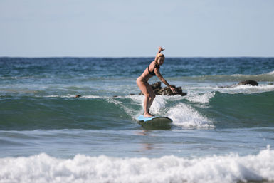 Female surfer balancing on a blue longboard, riding a small turquoise ocean wave toward a rocky shoreline under a clear sky — beach surfing scene