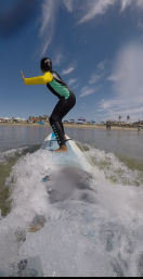 Person in a yellow-and-green wetsuit balancing on a surfboard through foamy shore break, heading toward a sandy beach with umbrellas, palm trees and blue skies — coastal surfing scene.