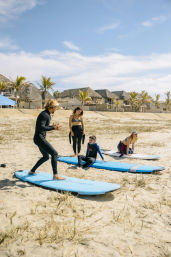 Surf instructor demonstrating stance on a blue foam board during a beach lesson while three students on surfboards watch on a sandy shore with palm trees and thatched beach huts under a bright blue sky.