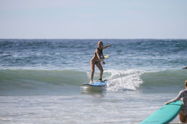 Surfer catching a gentle ocean wave on a blue surfboard near a sunny sandy beach.
