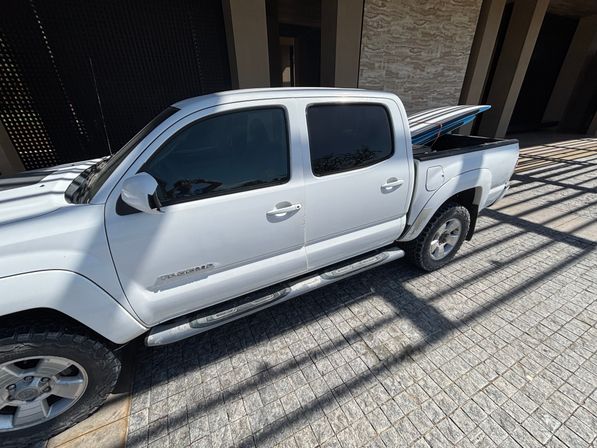 White crew-cab pickup truck parked on a sunlit tiled driveway with a surfboard in the bed, tinted windows, side steps and pergola shadow pattern — beach-ready vibe.