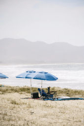 Sunny sandy beach with two blue umbrellas shading folding chairs and surfboards by the shoreline, calm ocean waves and hazy coastal mountains beyond.