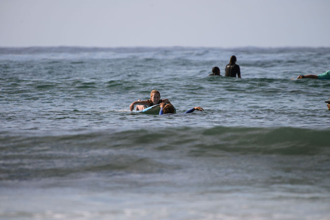 Surfers paddling on surfboards in the calm blue ocean with gentle rolling waves and a distant horizon, coastal beach scene.