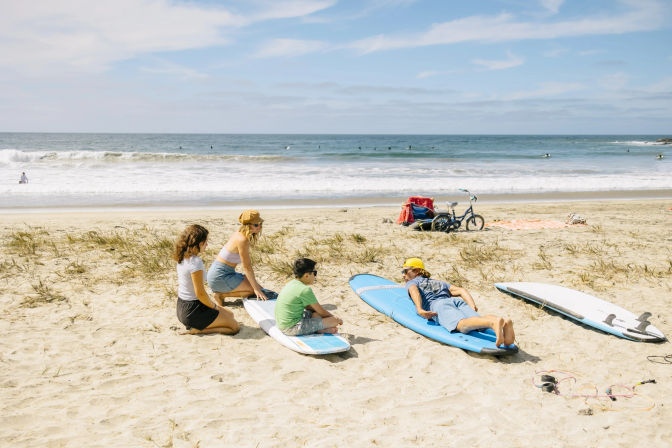 Sunny sandy coastline surf lesson with an instructor on a blue surfboard and three students seated on boards near the shoreline, ocean waves and surfers in the background.