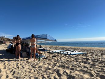 Sunny beach scene: group of people gathered under a blue umbrella on a sandy shore with several surfboards lined up near the calm ocean and clear blue sky.
