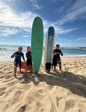 Four surfers on a sunny sandy beach holding tall green and blue surfboards at the shoreline with ocean waves and a blue sky, ready to ride the waves.