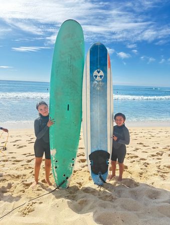 Two kids in wetsuits posing with tall turquoise and blue surfboards planted upright in the sand on a sunny beach with gentle ocean waves and a bright blue sky.