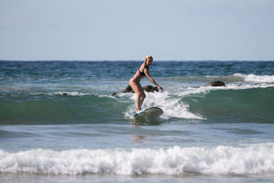 Female surfer in a black bikini riding a small turquoise wave toward a sunny coastal beach with rocks and a calm ocean horizon.