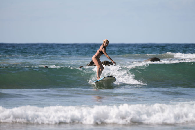Female surfer in a black bikini riding a small turquoise wave toward a sunny coastal beach with rocks and a calm ocean horizon.