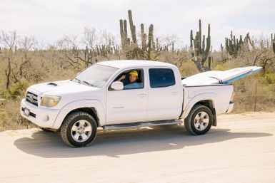 White pickup truck on a sandy desert road with a surfboard in the bed and a smiling driver waving from the open window in front of tall cacti