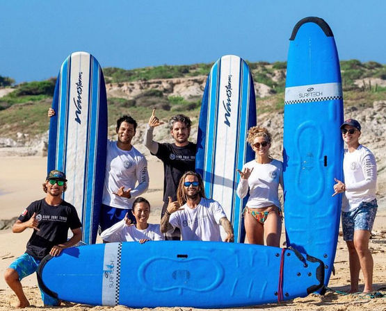 Group of surfers posing on a sunny sandy beach with bright blue longboard surfboards, smiling and flashing shaka signs in front of coastal dunes and a clear blue sky — surf lesson vibe.