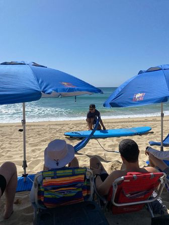 Sunny oceanfront beach scene with blue striped umbrellas and chairs, people relaxing on the sand watching a surf instructor kneel on a blue surfboard near gentle waves under a clear sky.