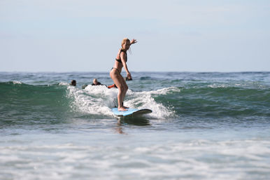 Female surfer in a black bikini standing on a blue longboard, riding a small ocean wave near shore with other surfers in the background on a sunny day.