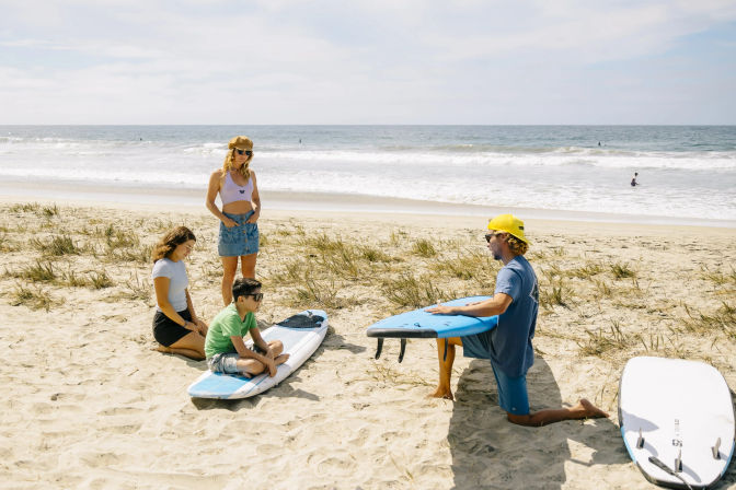 Surf instructor kneeling with a blue surfboard teaching a small group—two adults and a child on surfboards—on a sunny sandy beach with gentle ocean waves in the background.