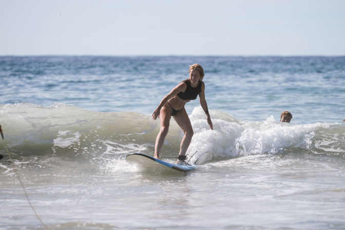 Surfer riding a small breaking wave on a blue surfboard at a sunny ocean beach, splashing water with a swimmer nearby