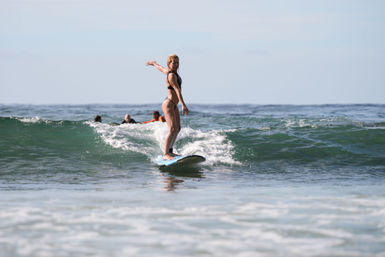 Surfer riding a small ocean wave on a blue longboard near shore, balanced with an outstretched arm and other surfers paddling in the background.