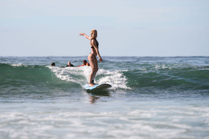 Surfer riding a small ocean wave on a blue longboard near shore, balanced with an outstretched arm and other surfers paddling in the background.