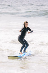 Smiling surfer in a black wetsuit balancing on a blue surfboard while riding a small wave along a sunny ocean beach