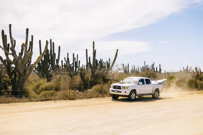 White pickup truck kicking up dust on a sandy desert road with a surfboard in the bed, surrounded by tall columnar cacti under a bright blue sky