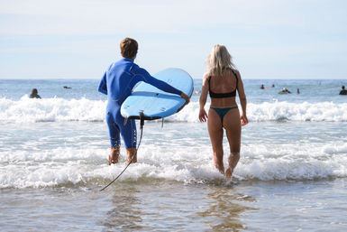 Two surfers wading into the ocean at a sunny beach — one in a blue wetsuit carrying a blue surfboard, the other in a bikini stepping into small waves with distant surfers beyond.