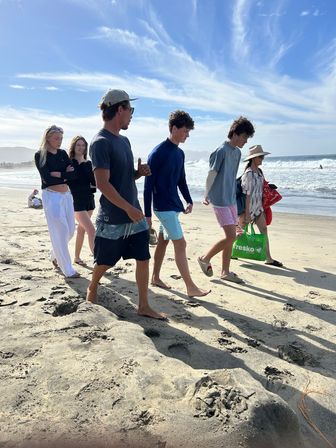 Six people walking along a sunny sandy beach by rolling ocean waves, dressed in casual summer clothes and carrying beach bags, casting long shadows on rippled sand under a blue sky with wispy clouds.