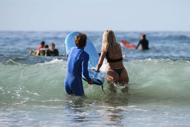 Two people wading into ocean waves at a sunny beach — one in a blue wetsuit holding a blue surfboard and a blonde woman in a bikini, with other surfers paddling in the background.